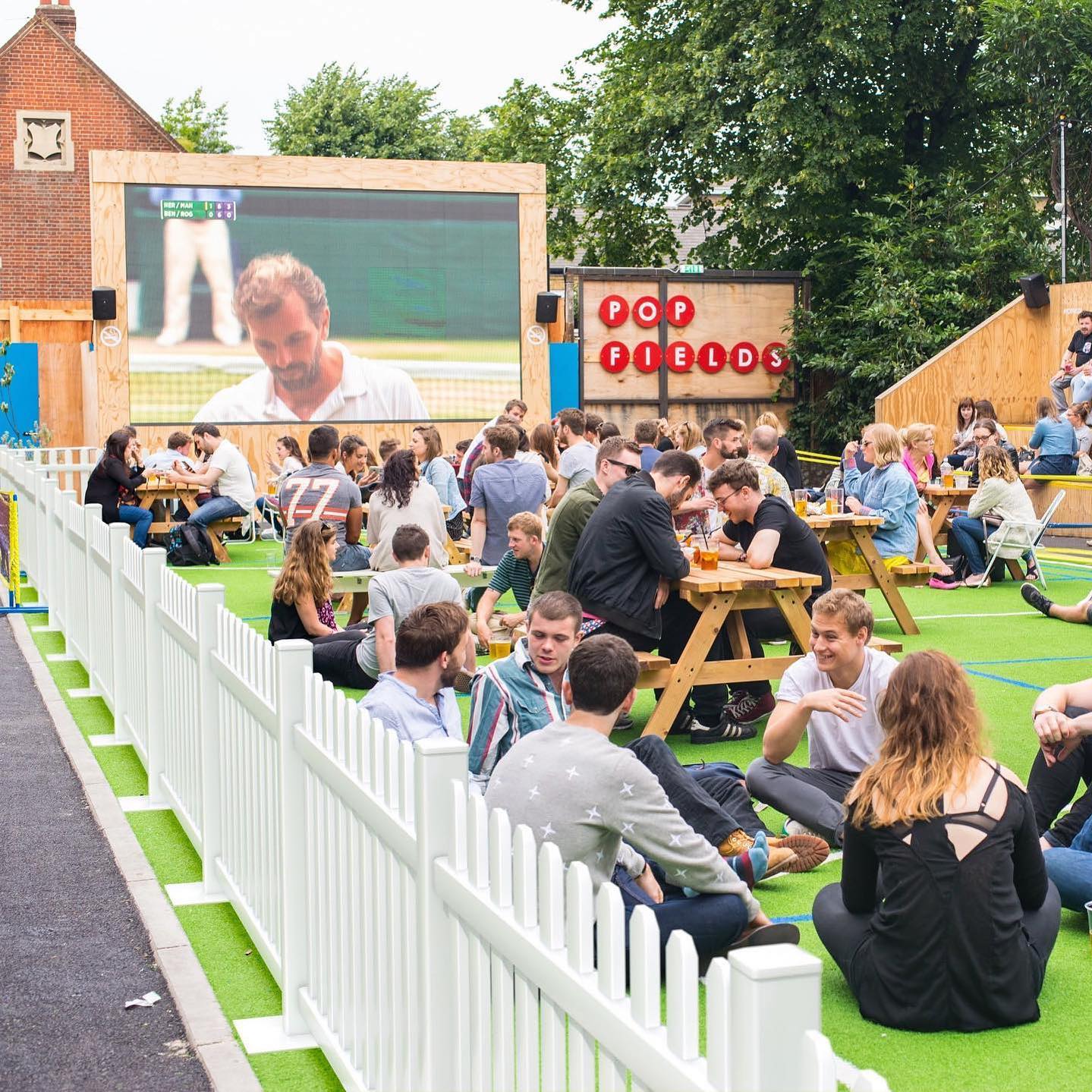 Outdoor social gathering at Pop Fields, featuring a large screen and picnic tables on green turf.