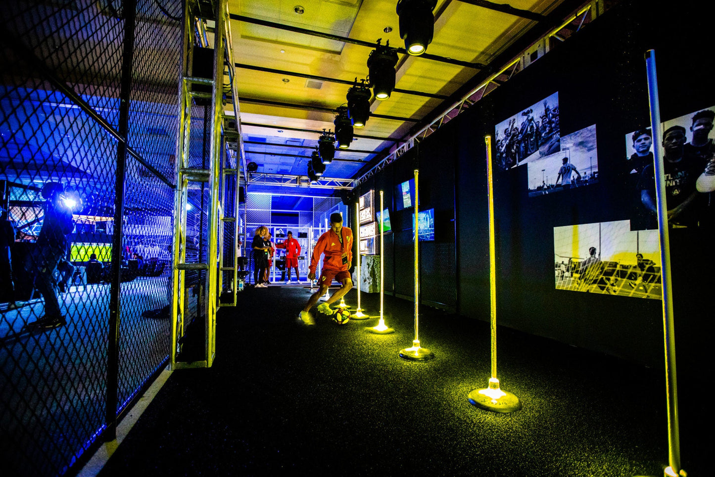 Indoor soccer training area featuring yellow training poles and a player dribbling a ball on synthetic turf.