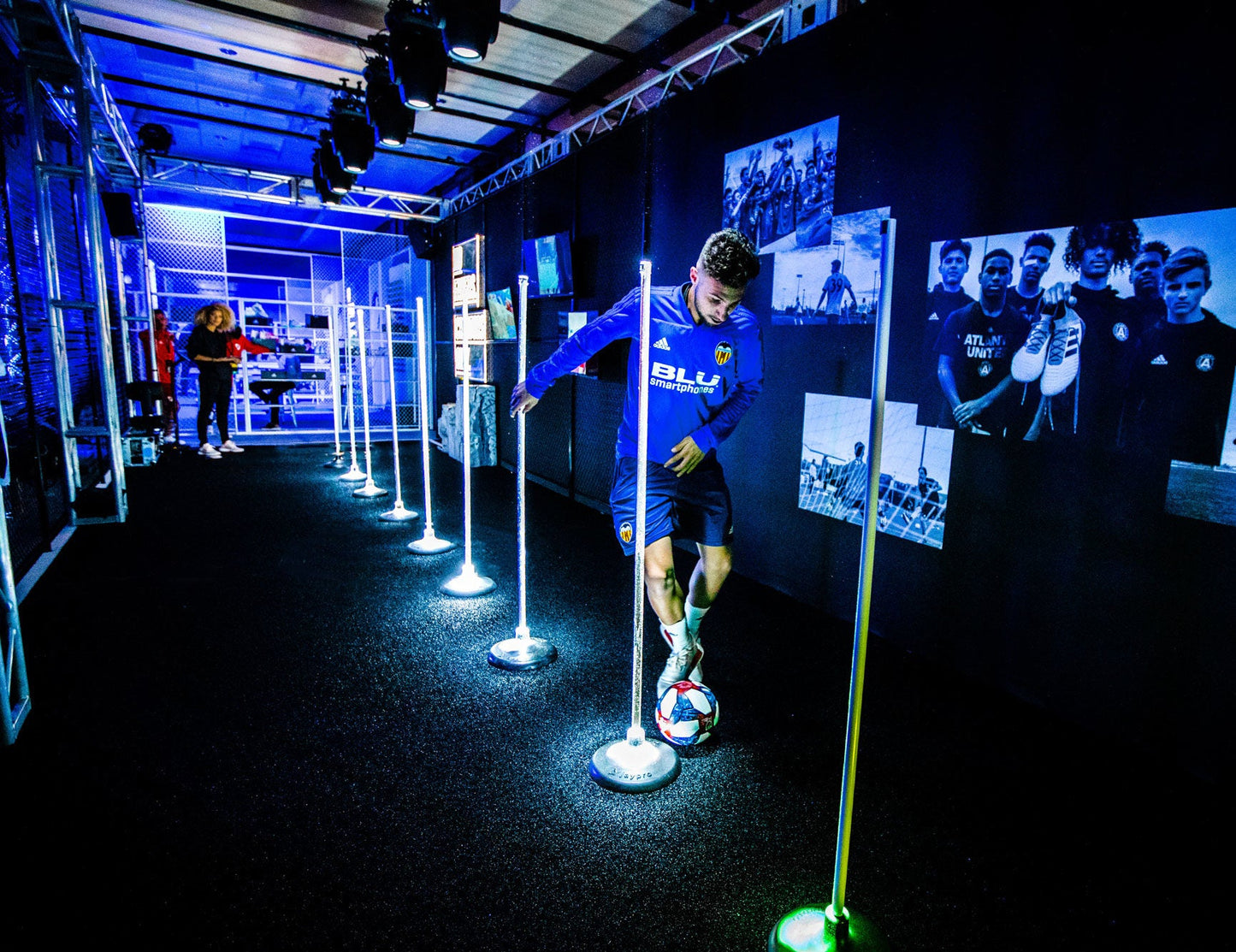 Male soccer player in blue jersey dribbling a ball on turf in a modern training setup with lights.