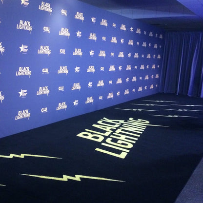 Blue backdrop and black carpet with 'Black Lightning' branding at a promotional event.