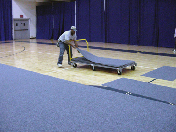 Man rolling out gray gym flooring on a cart in a gymnasium setting.