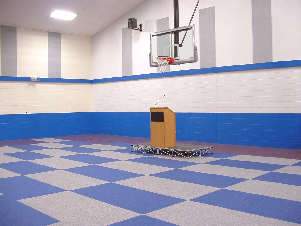 Indoor basketball court featuring blue and grey tiled flooring, with a podium on a raised platform.