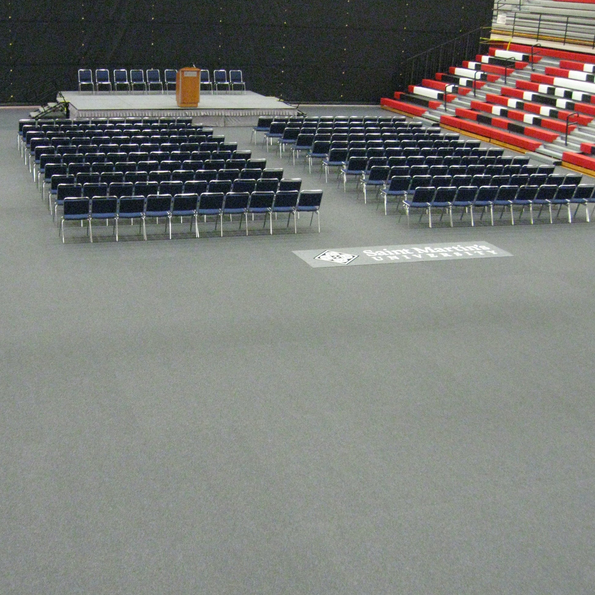 Arranged blue chairs on gray floor for an event, with a podium on a raised platform in a gymnasium.
