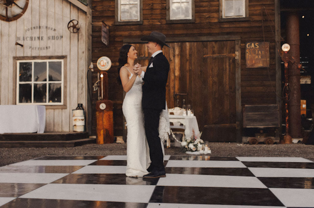 Couple in stylish wedding attire dancing on a checkerboard floor in a rustic outdoor setting.