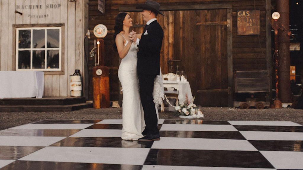 Couple dancing on a black and white checkerboard floor at a rustic outdoor wedding venue.