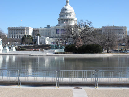 View of the US Capitol building with reflective water and security barriers in the foreground.