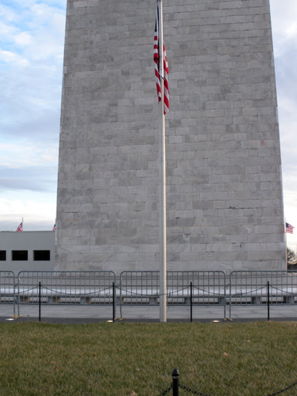 Washington Monument base with grass, flagpole, and American flags waving on a cloudy day.