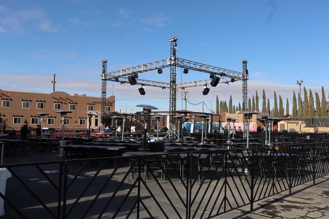 Outdoor event setup with a metal stage, black tables, and heating lamps under a clear blue sky.