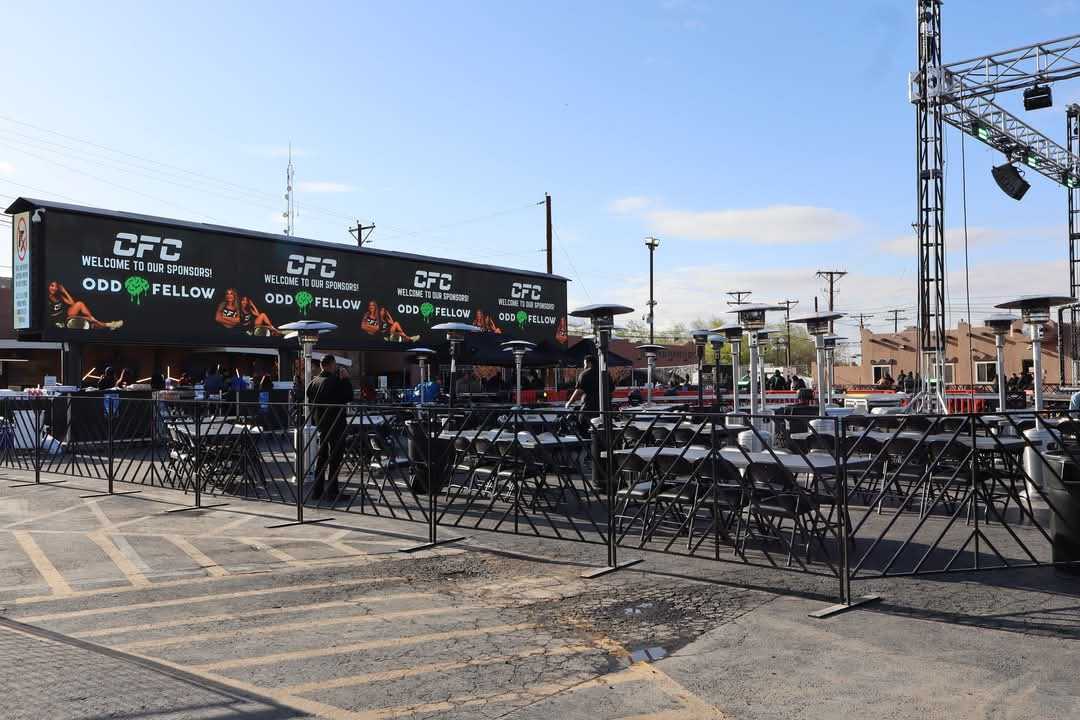 Outdoor event setup with black folding chairs and heating lamps, featuring large sponsor banners.