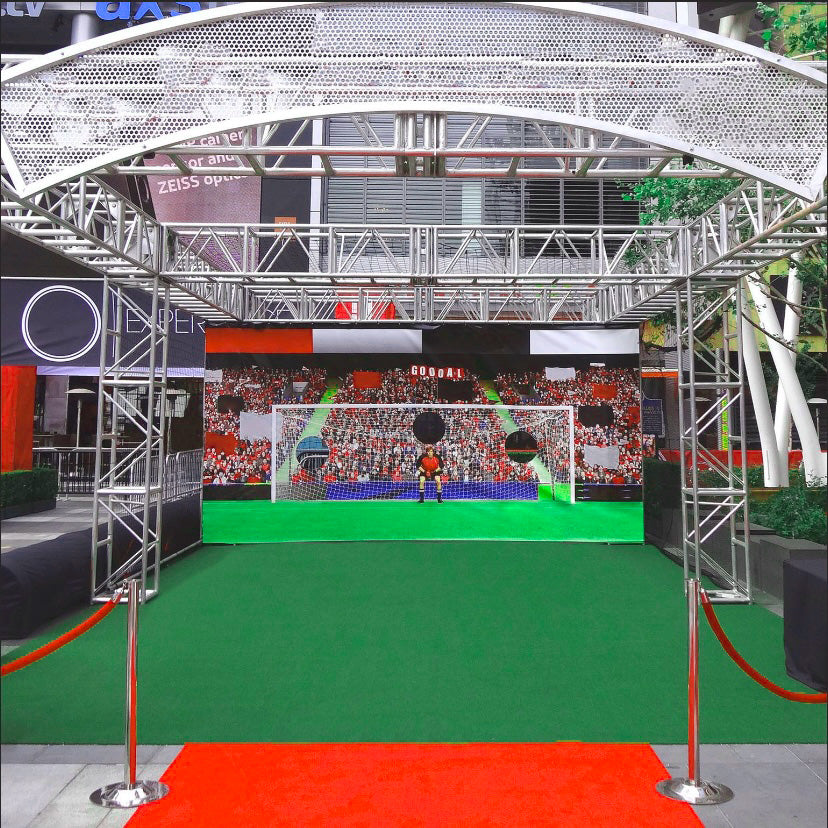 Interactive soccer goal setup with green turf, red carpet, and backdrop of cheering fans.