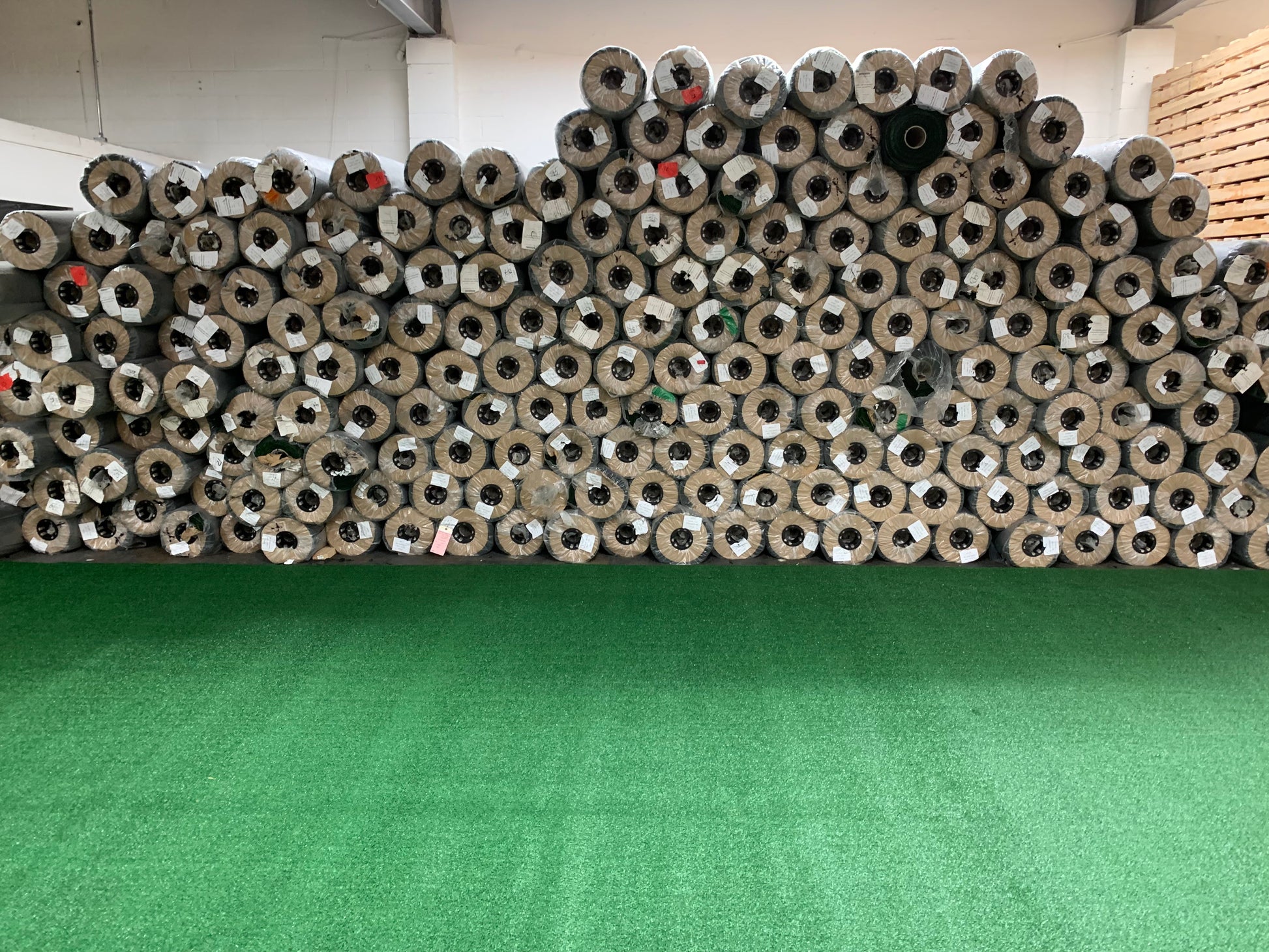 Stacked rolls of artificial turf in various shades of green, displayed in a warehouse setting.