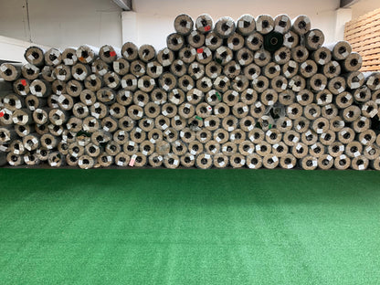 Stacked rolls of artificial turf in various shades of green, displayed in a warehouse setting.