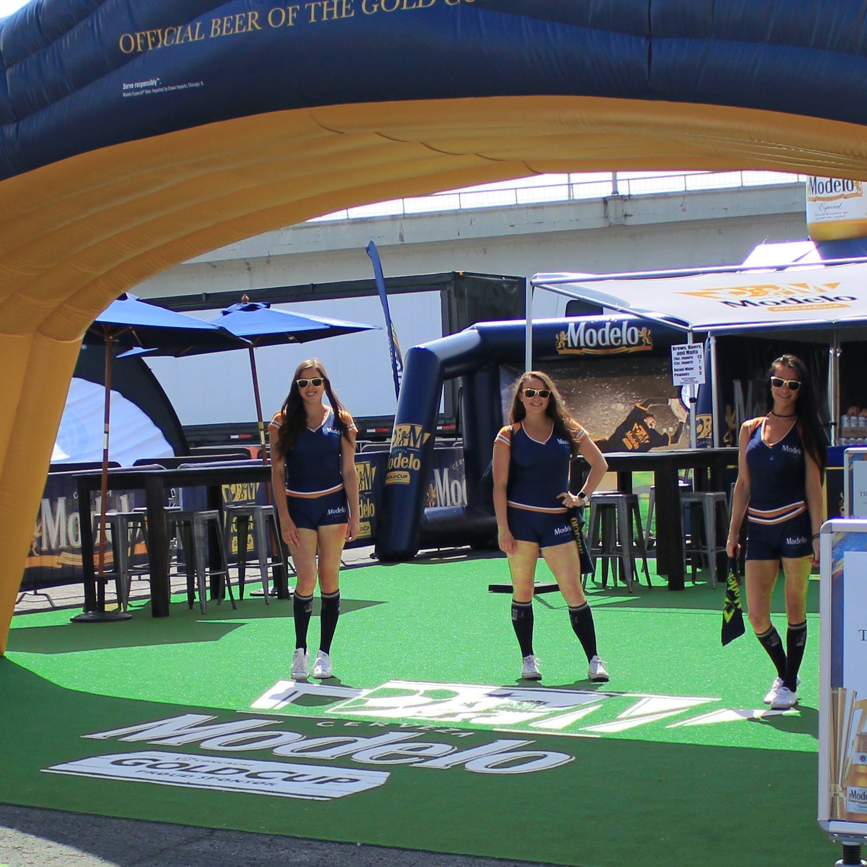 Three models in navy outfits promoting Modelo beer at a vibrant event entrance.