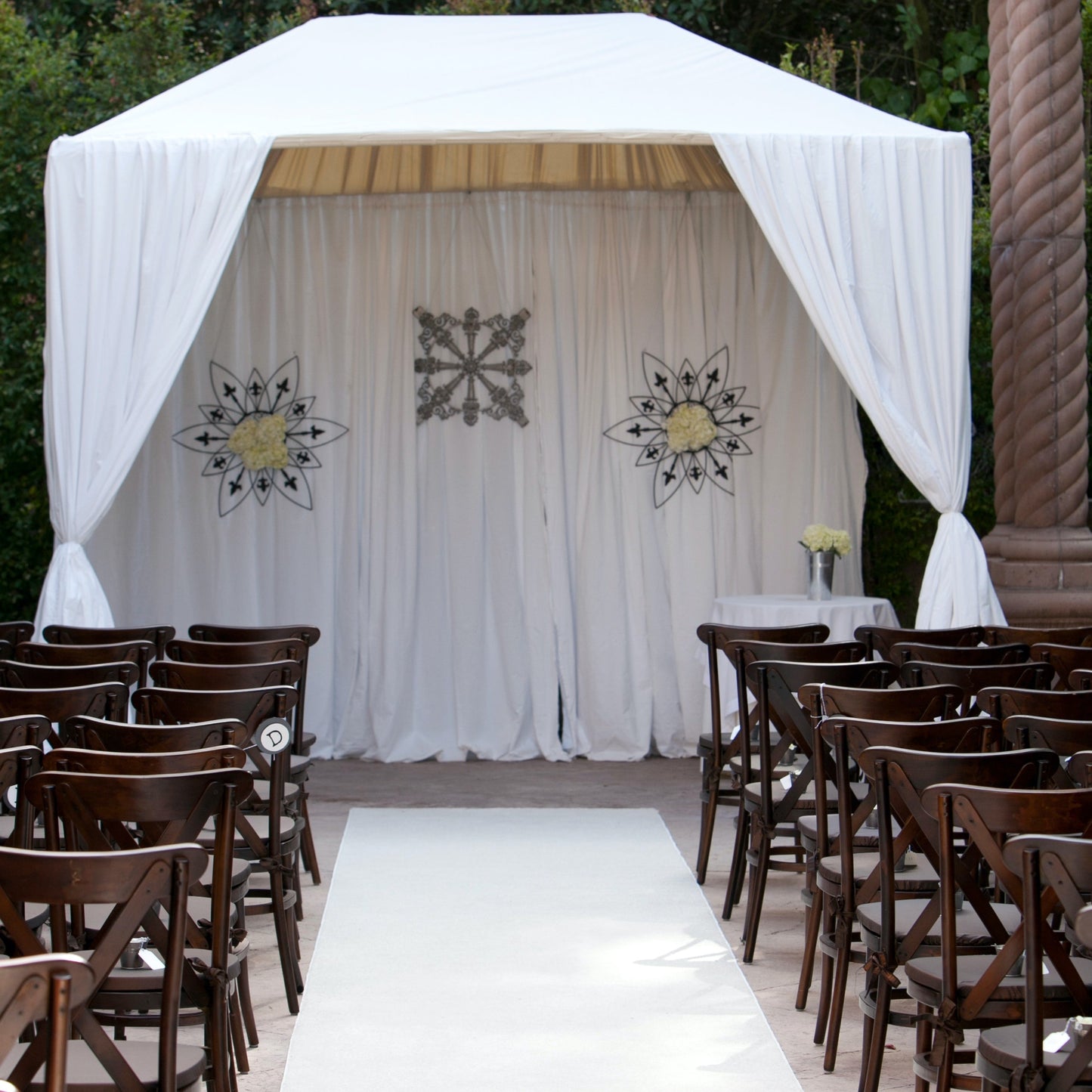 Elegant outdoor wedding altar with white drapes, floral decor, and wooden chairs for guests.