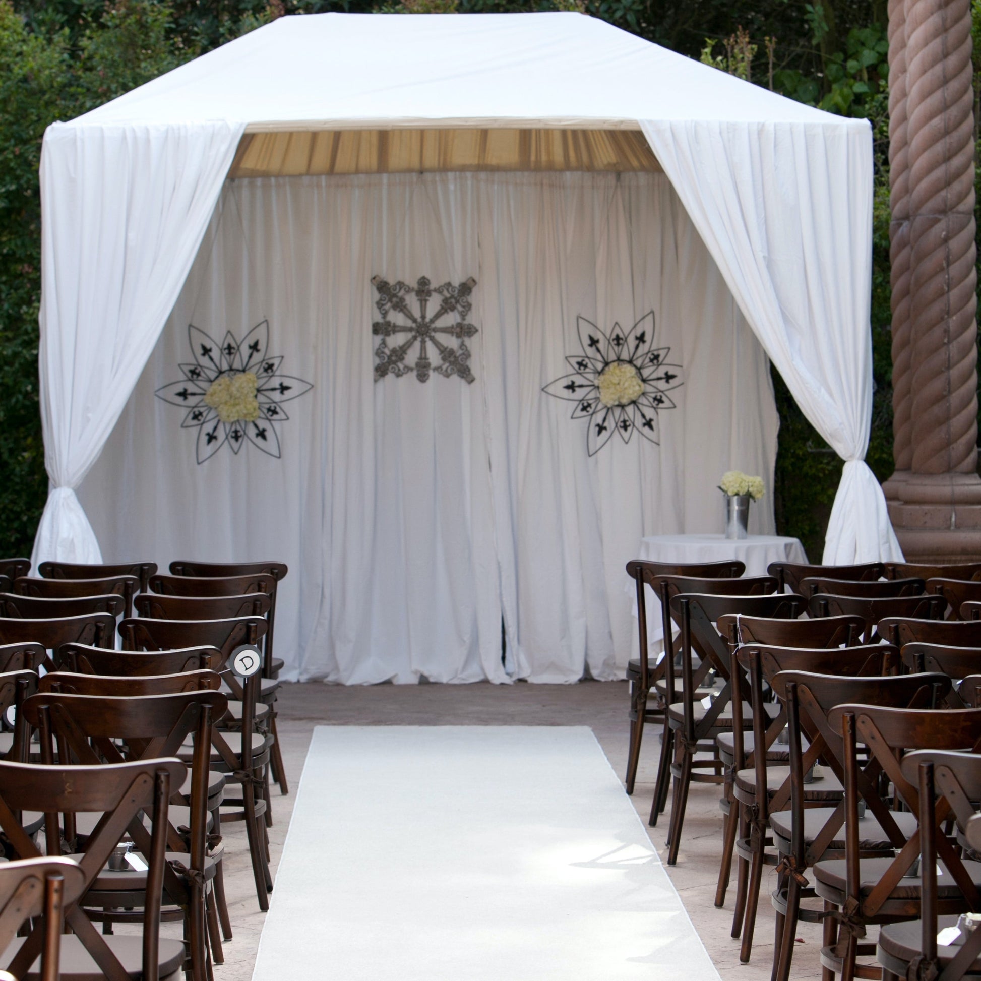 Elegant outdoor wedding altar with white drapes, floral decor, and wooden chairs for guests.
