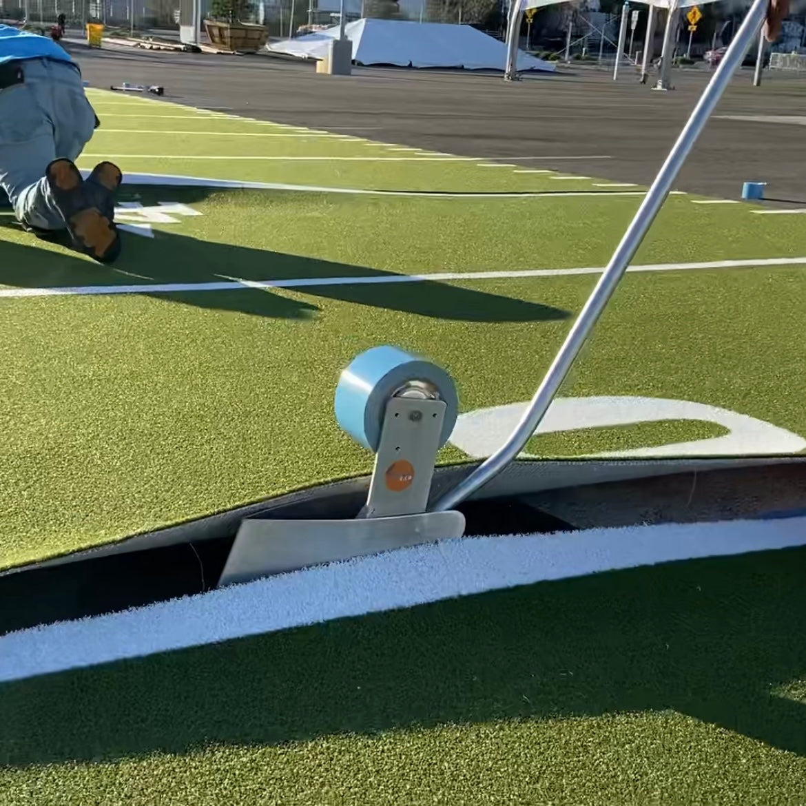 Grass field line marking tool in use, featuring a metal frame and blue roller on artificial turf.