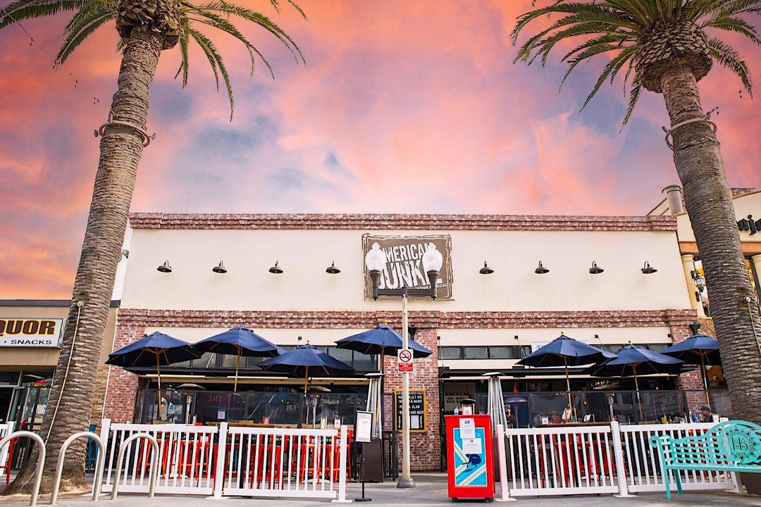 Exterior view of American Junk restaurant with blue umbrellas, red chairs, and palm trees at sunset.