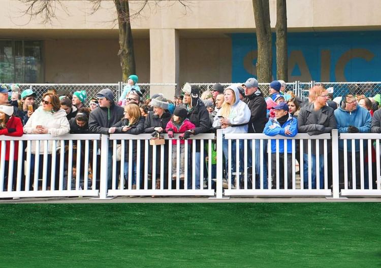 Crowd of people at an outdoor event, wearing winter jackets and hats, gathered behind a white fence.