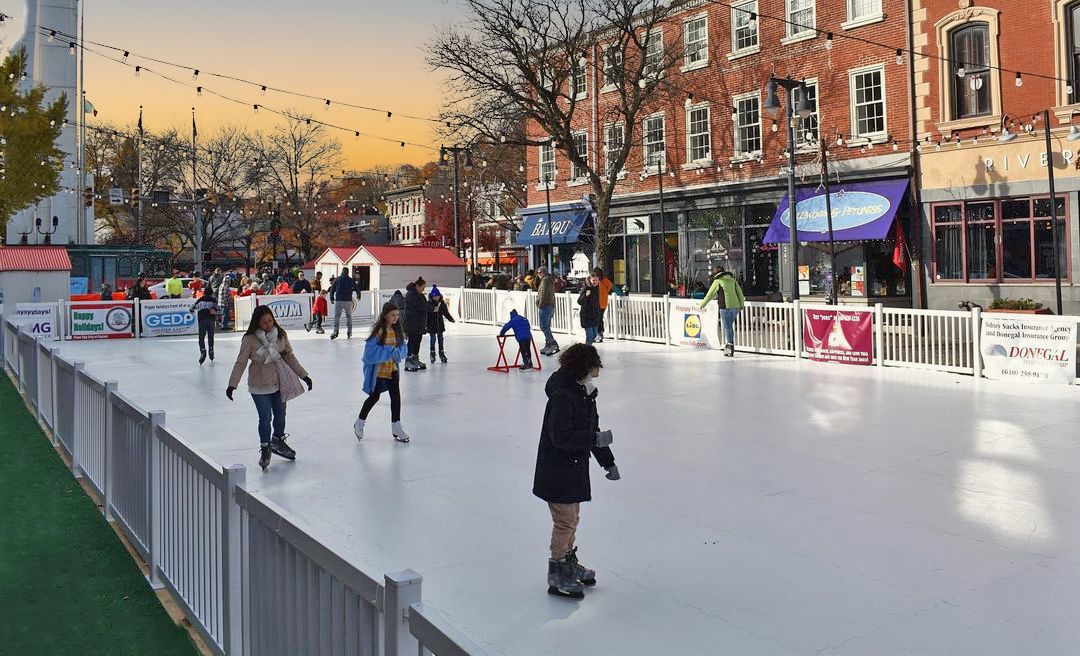 Outdoor ice skating rink with skaters, surrounded by vibrant autumn trees and shops, festive atmosphere.